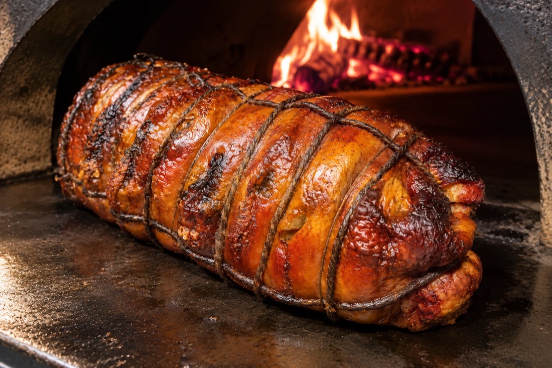 Porchetta legata e dorata appena uscita dal forno a legna, con la fiamma accesa sullo sfondo
